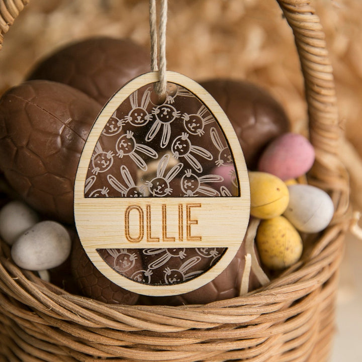 A wicker basket with chocolate and candy eggs, featuring a wooden Personalised Golden Egg Easter Tag adorned with bunny illustrations and the name OLLIE engraved on it.