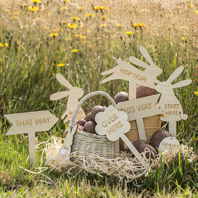 An arrangement of Easter eggs in baskets in a field. There are 6 Easter hunt signs in light bamboo. Three in the shape of bunnies saying start here, hop hop! and nearly there. Two signs that are arrow shaped saying this way and that way. One cloud shaped sign saying over here. 