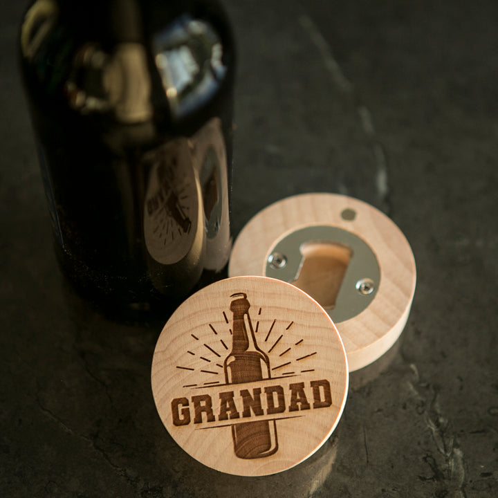 A Personalised Magnetic Bottle Opener (3 designs available), engraved with GRANDAD and a beer bottle motif, rests by a dark glass bottle on a sleek countertop—the ideal customised gift for men.