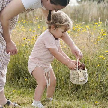 A young child puts brown eggs with Personalised Easter Bunny Tags into a wicker basket as an adult stands nearby in a grassy field with yellow wildflowers, getting ready for the Easter egg hunt.