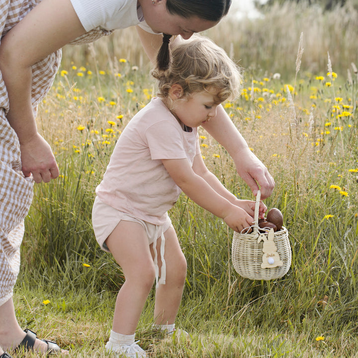 A young child puts brown eggs with Personalised Easter Bunny Tags into a wicker basket as an adult stands nearby in a grassy field with yellow wildflowers, getting ready for the Easter egg hunt.