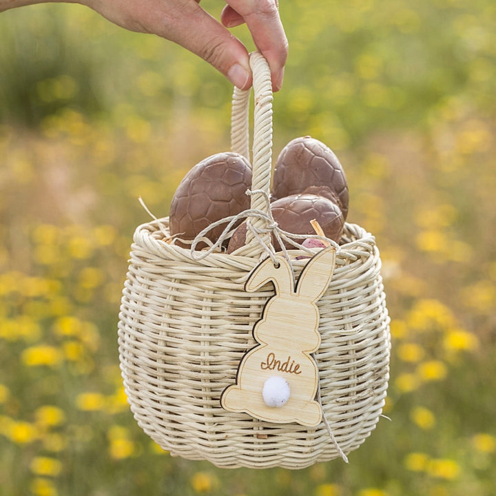 A hand holds a woven basket with a Personalised Easter Bunny Tag and chocolate eggs, set outdoors in a field of yellow flowers.