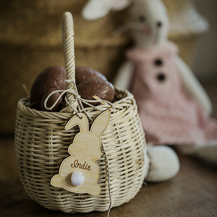 A wicker basket holding two chocolate eggs and a Personalised Easter Bunny Tag labelled "Indie" sits in front of a pink-dressed stuffed rabbit toy, with a softly blurred background.