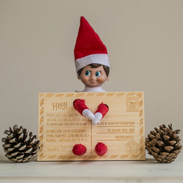 The Elf on the Shelf Arrival Postcard features a festive elf doll in a red hat holding a personalised postcard, with two pinecones beside it—a charming addition to your Christmas range.