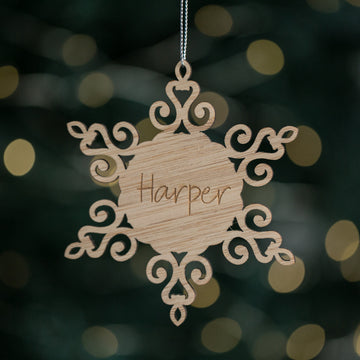 The Personalised Christmas Snowflakes ornament features intricate wooden cut-out designs and hangs from a silver string. "Harper" is engraved in the center, creating a unique custom piece against a backdrop of festive, blurred holiday lights.