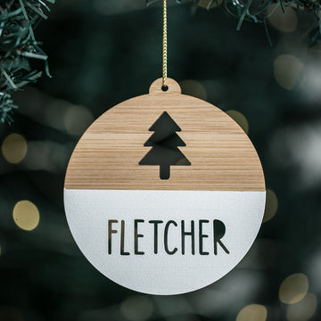 A personalized Christmas tree bauble with a wooden texture, light bamboo background, and a black and white pine tree design in the center. The name &