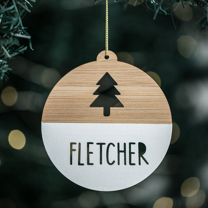 A personalized Christmas tree bauble with a wooden texture, light bamboo background, and a black and white pine tree design in the center. The name &