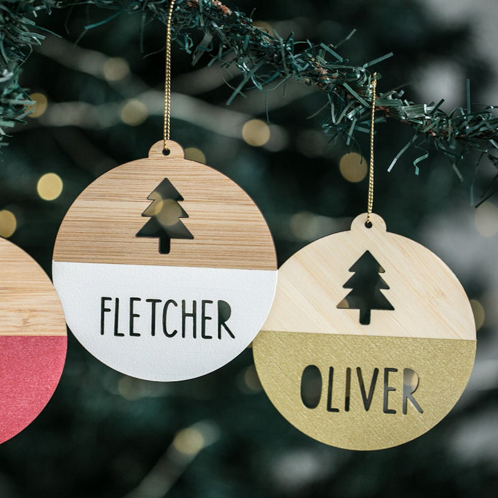 Two Personalised Christmas Tree Baubles hang on a branch, each featuring a cut-out tree and customised with names in capitals, finished in white and gold. Softly blurred festive lights shimmer in the background.