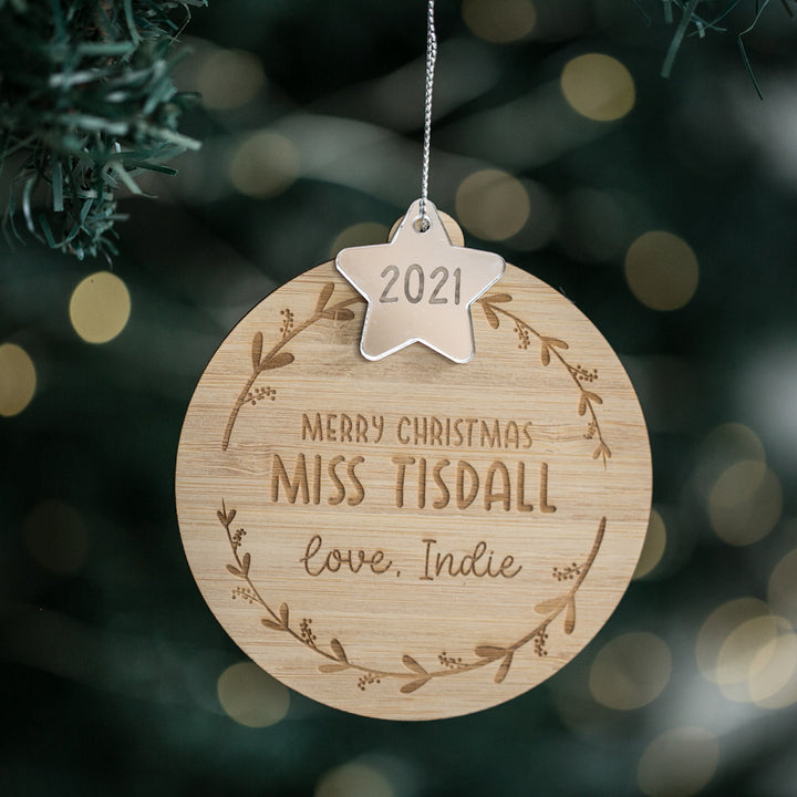 The Star Teacher Christmas Bauble, engraved with “Merry Christmas Miss Tisdall love, Indie” and topped with a 2021 metal star, hangs on a tree—an ideal personalised teacher gift with blurred festive lights in the background.