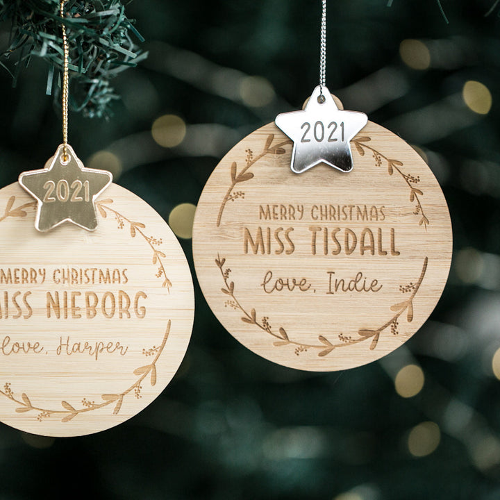 Two Star Teacher Christmas Baubles hang on a tree, each engraved with "Merry Christmas" and the names: MISS NIEBORG, love, Harper and MISS TISDALL, love, Indie. Both ornaments feature a 2021 star charm.
