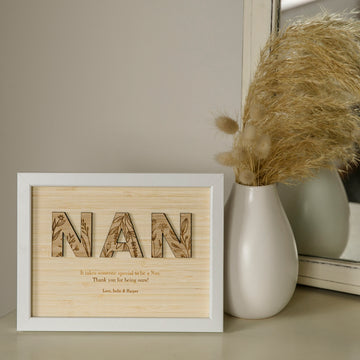 A Wildflower Frame Insert (frame not included) with “NAN” and floral designs sits by a white vase of pampas grass in front of a mirror—a unique engraved wildflower gift to thank Nan.