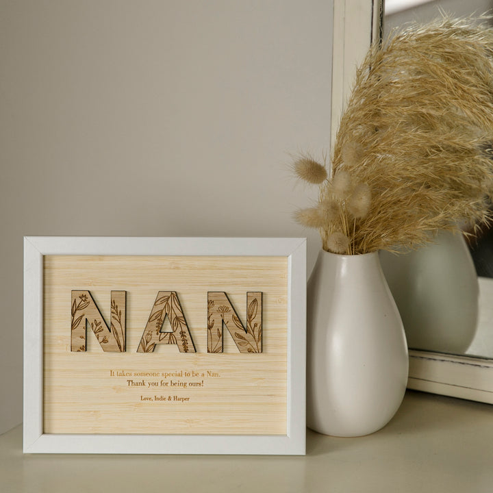 A Wildflower Frame Insert (frame not included) with “NAN” and floral designs sits by a white vase of pampas grass in front of a mirror—a unique engraved wildflower gift to thank Nan.