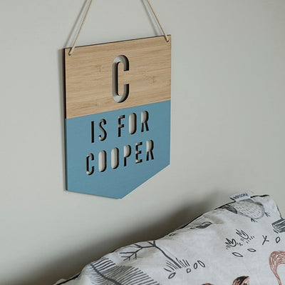 A Personalised Alpha Banner hangs on the wall, reading C IS FOR COOPER with its lower half painted blue. Below, a white pillow or bedding with animal illustrations.