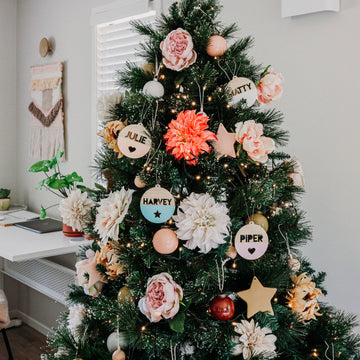 A decorated Christmas tree with large flowers, string lights, and The Original Personalised Christmas Bauble for each name—Julie, Natty, Harvey, and Piper—stands in a modern, cozy room with neutral walls.