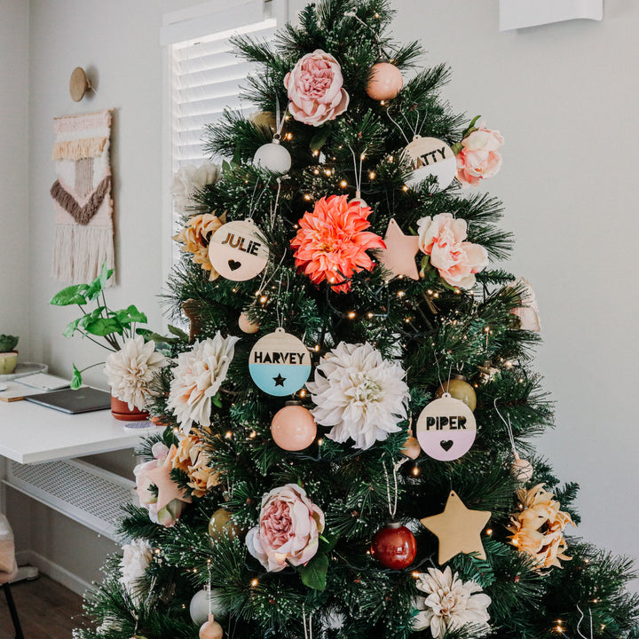 A decorated Christmas tree with large flowers, string lights, and The Original Personalised Christmas Bauble for each name—Julie, Natty, Harvey, and Piper—stands in a modern, cozy room with neutral walls.