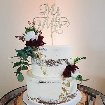 A light bamboo cake topper saying Mr and Mrs, sitting on a white cake with a floral arrangement.