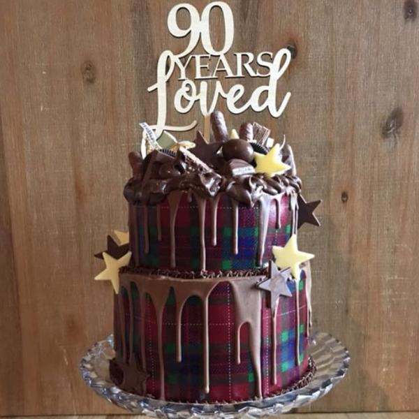 A two-tier chocolate cake with a plaid pattern, chocolate drip, star decorations, and the Years Loved Personalised Cake Topper reading "90 Years Loved," displayed on a glass stand against a wooden background.