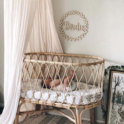 A leafy-look bamboo wreath, surrounding a bamboo personalised wall name saying Birdie. Hanging above a bassinet with a baby inside.