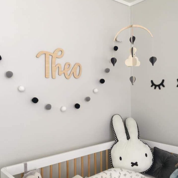 A neutral, minimalist baby nursery features a "Theo" Lovely Lettering Handlettered Wall Name in wooden letters, a black, white, and gray pom-pom garland, bunny and star pillows in a white crib, and a cloud mobile above.