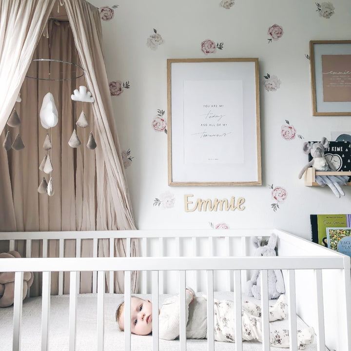 A baby lies in a white crib in a pastel nursery with floral wallpaper, wall art, cloud mobile, and a Lovely Lettering Handlettered Wall Name sign reading "Emmie" above the crib.