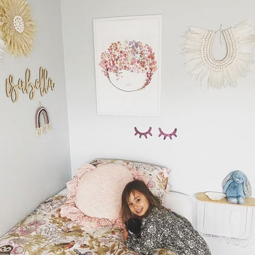 A bamboo wall name saying Isabella, hanging in a little girls room with a girl sitting on her bed.