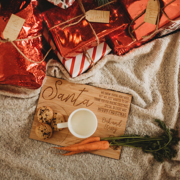 The Personalised Santa Treat Board holds a glass of milk, two cookies, and two carrots. Placed on a beige blanket, it’s surrounded by wrapped Christmas presents in red and white festive paper.