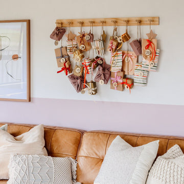 A wooden rack above a tan leather couch displays 25 wrapped gifts, each labelled with the Advent Calendar Tags Set. Pillows decorate the couch below.