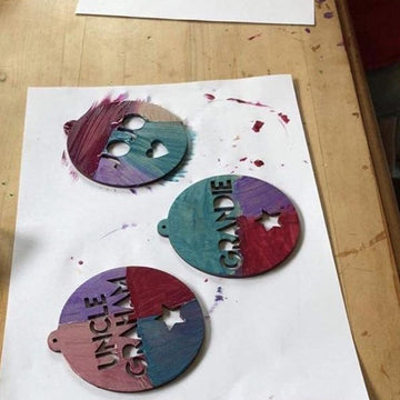 Three Decorate Your Own Christmas Bauble ornaments with cut-out names (UNCLE GRAHAM, GRANDIE) rest on white paper atop a wooden table. These colorful DIY baubles are a perfect family Christmas craft for your loved ones.