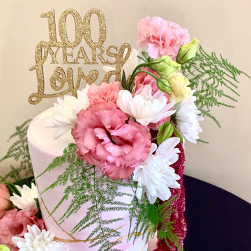 A light pink cake adorned with pink and white flowers, green ferns, and the Years Loved Personalised Cake Topper in gold glitter reading "100 Years Loved," set against a neutral background—perfect for celebrating special milestones.
