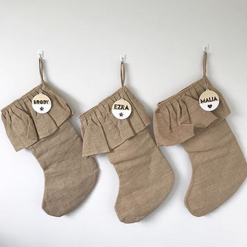 Three beige stockings hang from a white wall, each with The Original Personalised Christmas Bauble tag displaying the names Brody, Ezra, and Malia in bold letters. The stockings are finished with ruffled cuffs.