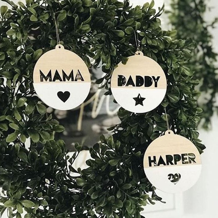 Three round baubles from The Original Personalised Christmas Bauble set hang on a wreath, labeled MAMA with a heart, DADDY with a star, and HARPER with two hearts—a charming family Christmas tradition.