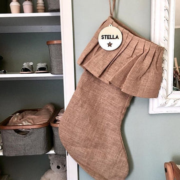 A burlap Christmas stocking hangs on the wall, decorated with The Original Personalised Christmas Bauble featuring a STELLA wooden tag and black star. Shelves nearby hold baskets, containers, and a white plush toy.