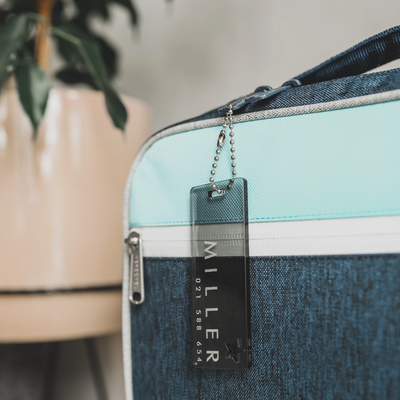 A close-up of The Details Key Ring—a blue and turquoise zipper bag accessory with a transparent tag labeled MILLER and a phone number—hangs from the zipper pull. A potted plant is blurred in the background, highlighting its practicality.