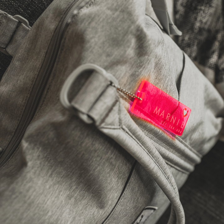 Close-up of a grey fabric bag with The Details Key Ring—a bright pink, translucent MARNI accessory—attached to the zipper. The bold key ring is in focus, while the bag appears slightly blurred in the background.