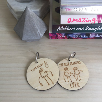 Two round light bamboo keyrings with a childs drawing etched on each one. One saying Nanny, the other saying The best Grandies ever. Sitting on a coffee table with books.