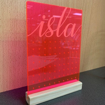 The Personalised Earring Holder is a neon pink acrylic sign with "isla" in cursive, standing upright in a wooden base. It features a grid of small holes for earrings and sits on a dark surface with a wooden background.
