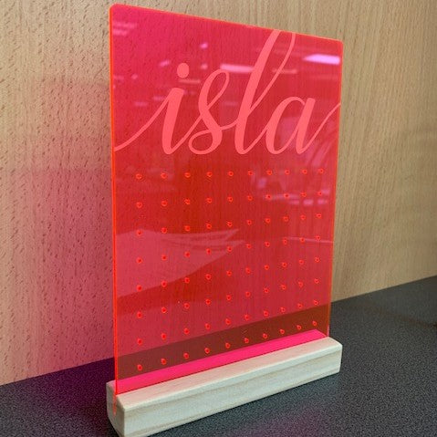 The Personalised Earring Holder is a neon pink acrylic sign with "isla" in cursive, standing upright in a wooden base. It features a grid of small holes for earrings and sits on a dark surface with a wooden background.