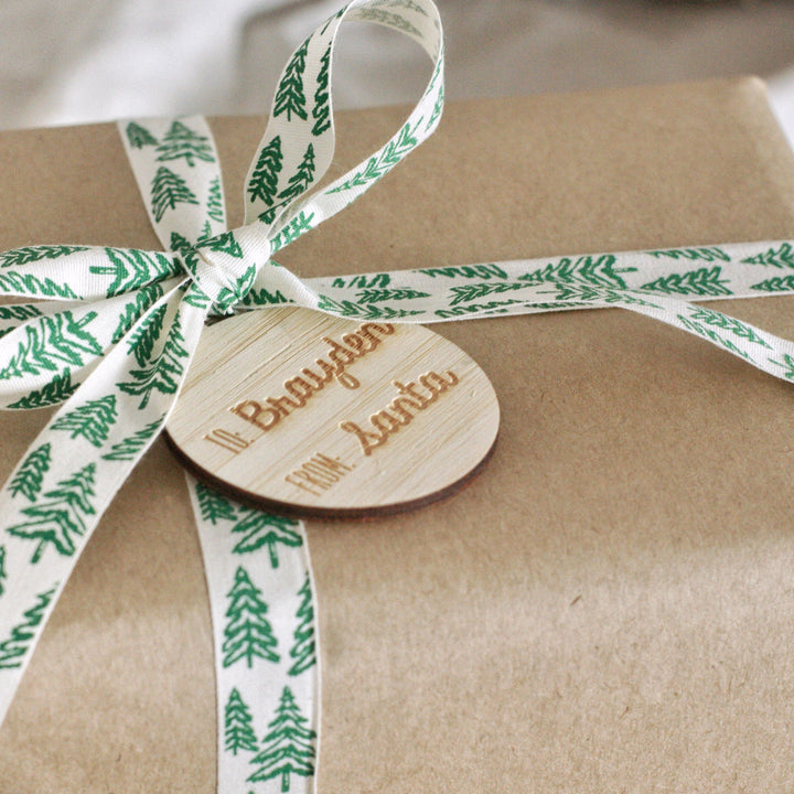 A gift wrapped in brown paper with a white ribbon featuring green pine trees, finished with a Personalised Christmas Gift Tag engraved "To: Brayden From: Santa".