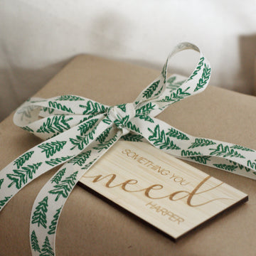 A gift wrapped in brown paper with a white ribbon printed with green pine branches, topped with a Personalised - Want, Need, Wear, Read Tag engraved with "Something you need" and "Harper" underneath.