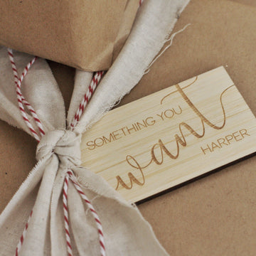 A Personalised - Want, Need, Wear, Read Tag engraved with "Something you want Harper" sits on a beige fabric-wrapped present tied with red and white string, set against brown paper packages.