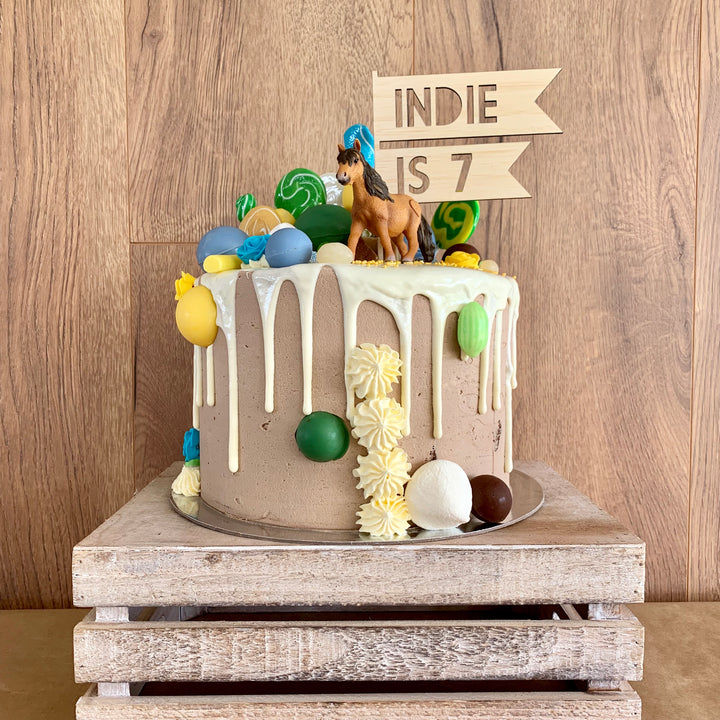 A flag shaped light bamboo cake topper saying Indie is 7. Sitting on a brown and white cake with a horse figurine, lollipops and lollies. 