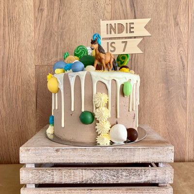 A flag shaped light bamboo cake topper saying Indie is 7. Sitting on a brown and white cake with a horse figurine, lollipops and lollies. 