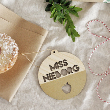 The Teacher Christmas Bauble, a round wooden ornament featuring MISS NIELBORG and an apple cutout, sits on beige fabric near festive decor—perfect as a personalised end-of-year gift for teachers.
