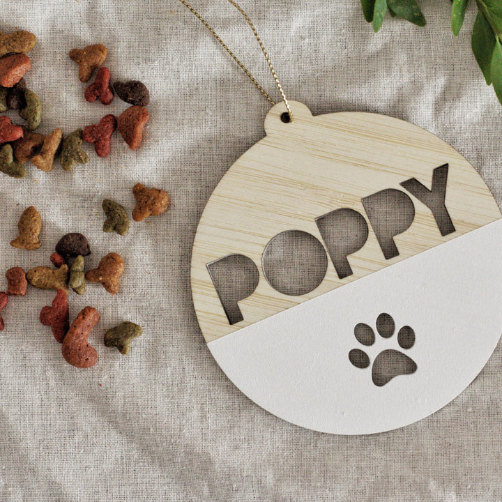 A Personalised Pet Christmas Bauble engraved with the name POPPY and a paw print rests on beige fabric next to multicolored dog treats and green leaves.