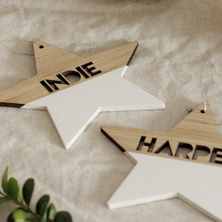Two Personalised Christmas Stars, star-shaped wooden ornaments with natural wood tops and white-painted bottoms, labeled INDIE and HARPE. Each has a hole and silver string for hanging—perfect festive decor.