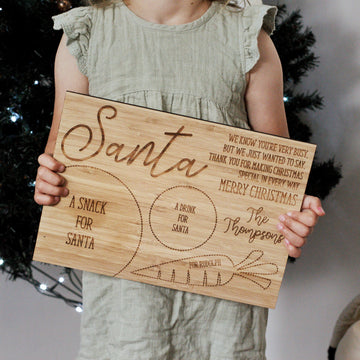 A child in a green dress holds a Personalised Santa Treat Board, custom-etched with sections for Santa’s snack, drink, and Rudolph’s treat. A festive engraved message and a Christmas tree in the background capture this charming holiday tradition.