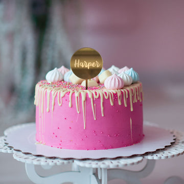 A round gold mirror cake topper saying Harper, on a pink and white drip cake with meringues. 