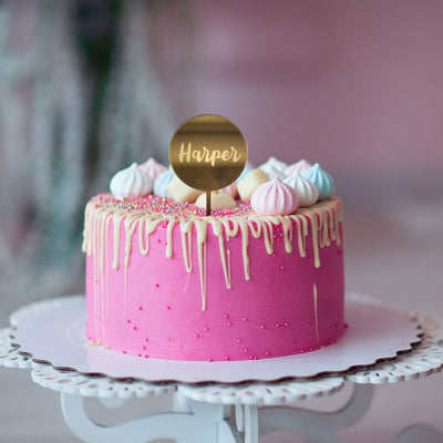 A round gold mirror cake topper saying Harper, on a pink and white drip cake with meringues. 