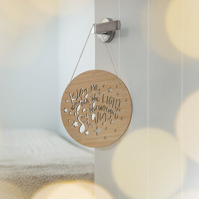 A round dark bamboo plaque saying Kiss me under the light of a thousand stars, with stars engraved. Hanging on a bedroom door. 
