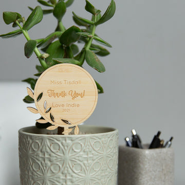 The Personalised Stem Teacher Planters feature a textured pot and a custom wooden plaque, making them an ideal teacher gift. In the background, a container holds pens, adding a practical touch to this thoughtful present.